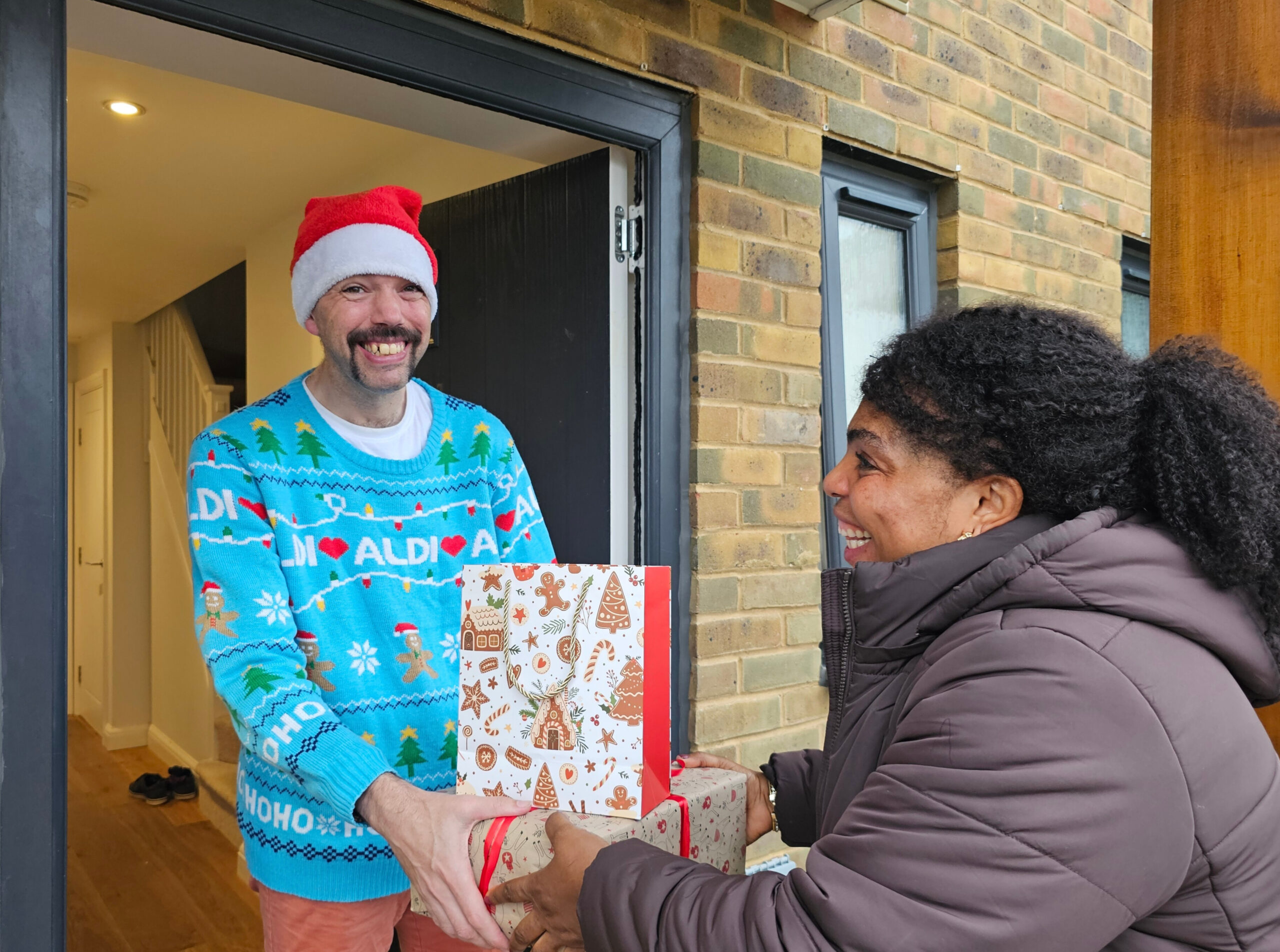 A man in a vibrant Christmas jumper receiving a present at his front door.