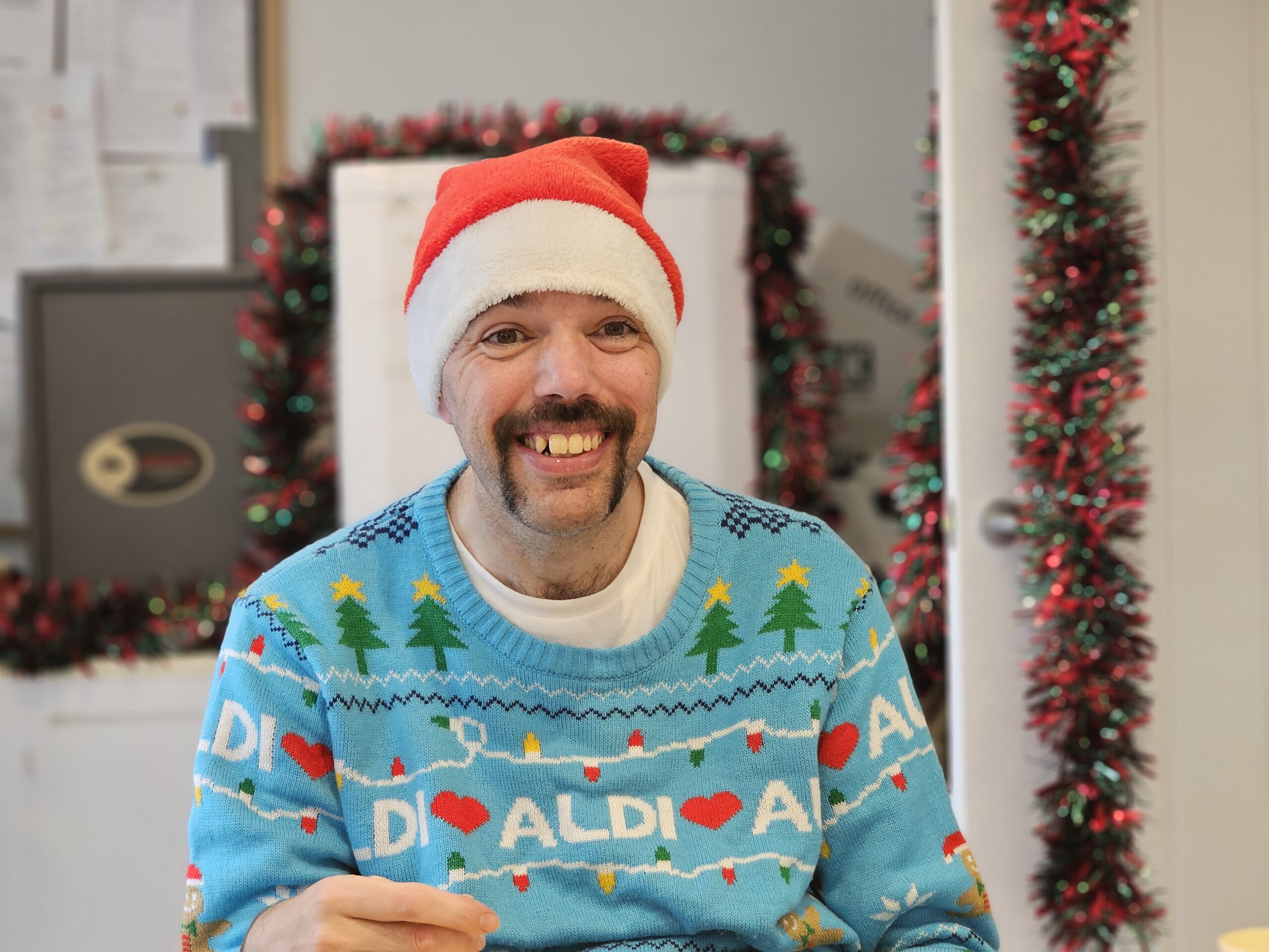 A man in a vibrant Christmas jumper making mince pies, smiling towards his helpful Support Worker.