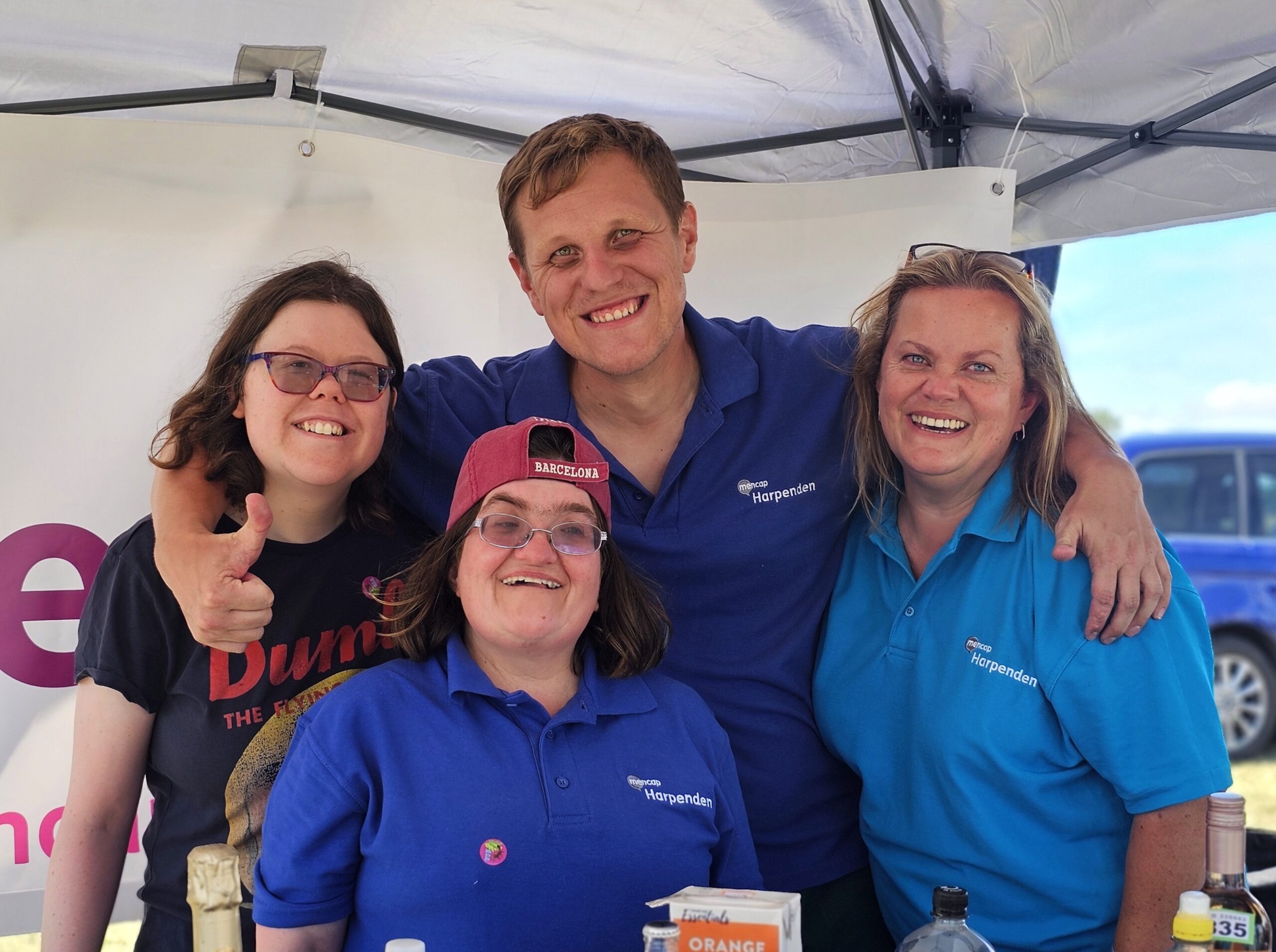 A group of four from Harpenden Mencap smiling at a fundraising stall, wearing branded blue polo shirts.
